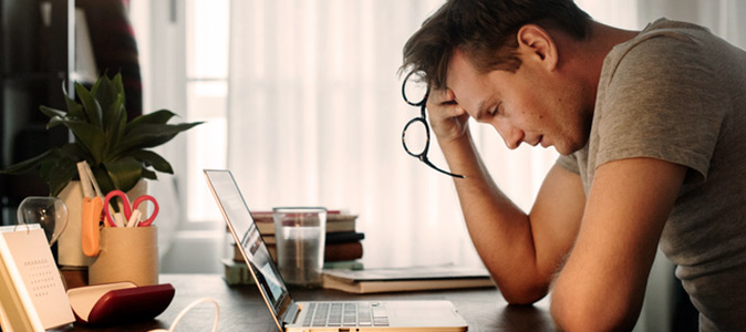 Homem sentado em frente a uma mesa de escritório, com a mão na cabeça e olhar cansado